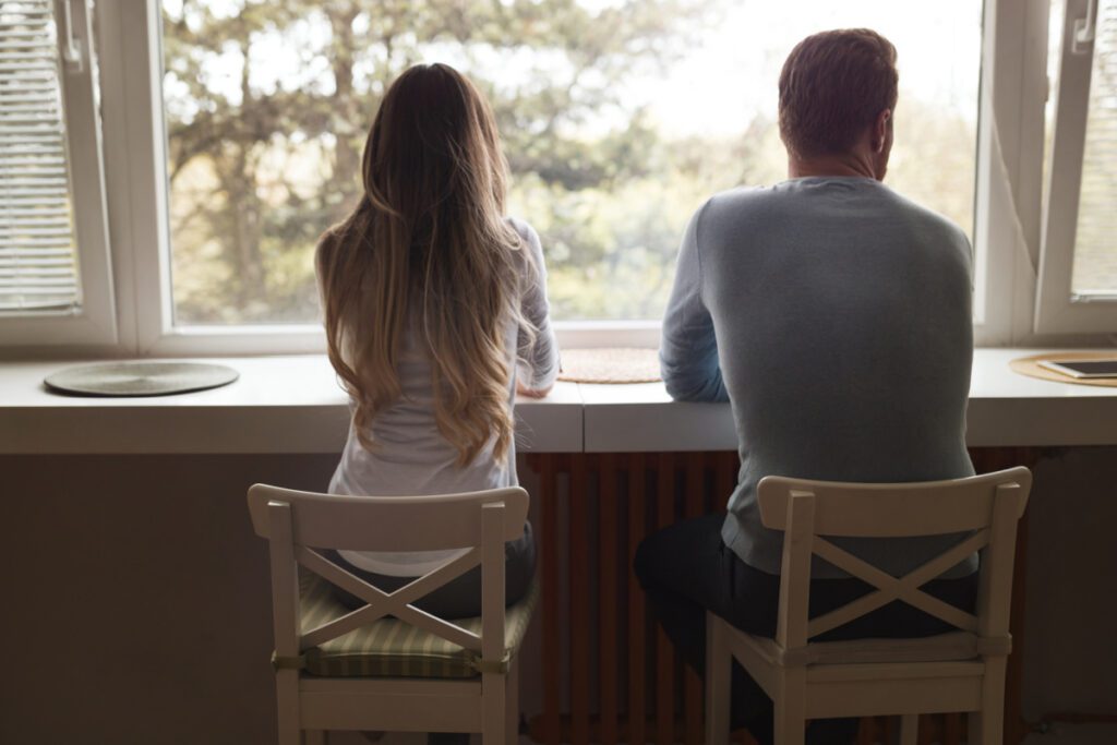 Couple in disagreement sitting with their backs to the camera, looking out the window, not speaking.