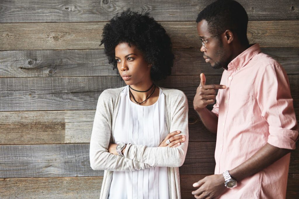 Woman with crossed arms standing beside man pointing finger to himself.