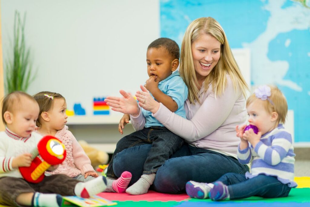 Blonde woman holding a toddler and playing with a group of babies at a daycare.