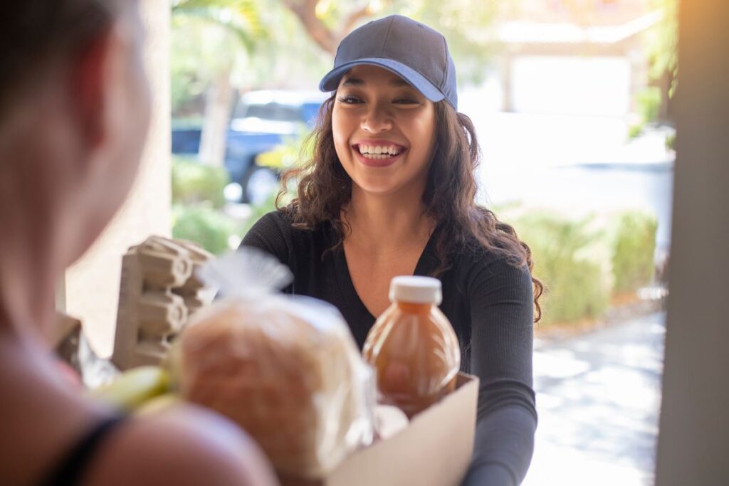 A young woman in a baseball cap delivering groceries to a person's home.