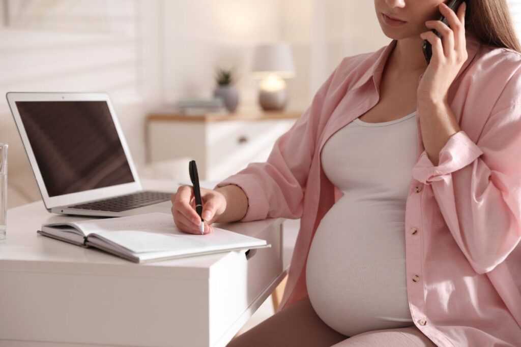 Pregnant woman writing in a notebook near her laptop while working on home