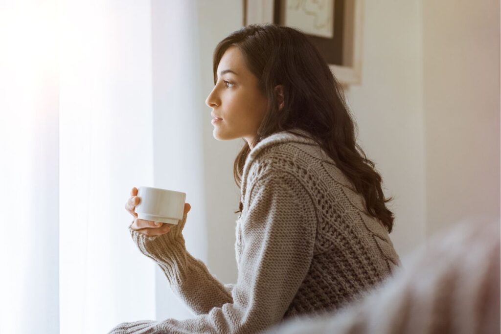 Woman looking out window with cup of tea, thinking.