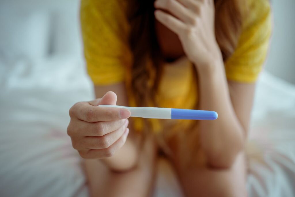 Teen girl in yellow shirt looking at pregnancy test while sitting on edge of her bed.