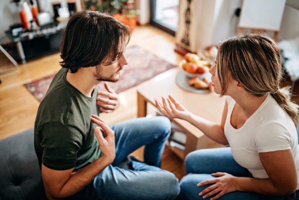 Woman and boyfriend arguing and gesturing with their hands while sitting in a living room.