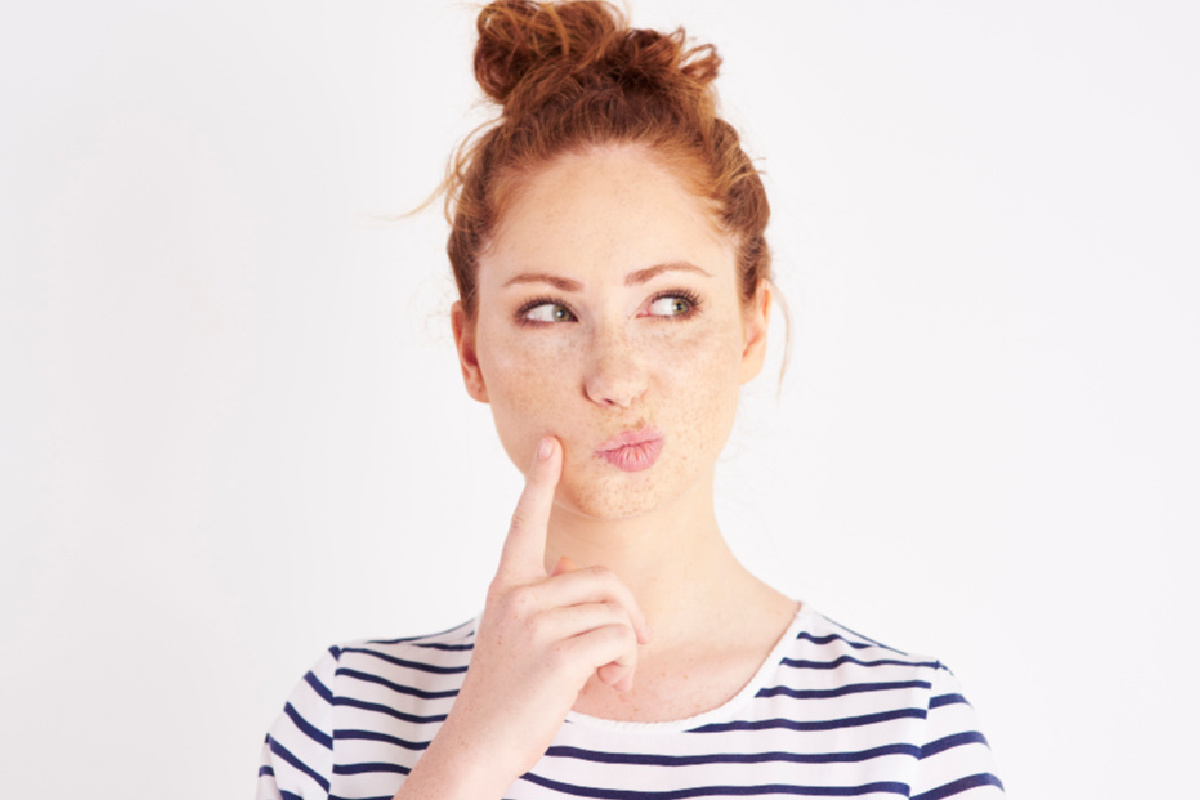 Red headed young woman with finger held to the side of her mouth, thinking