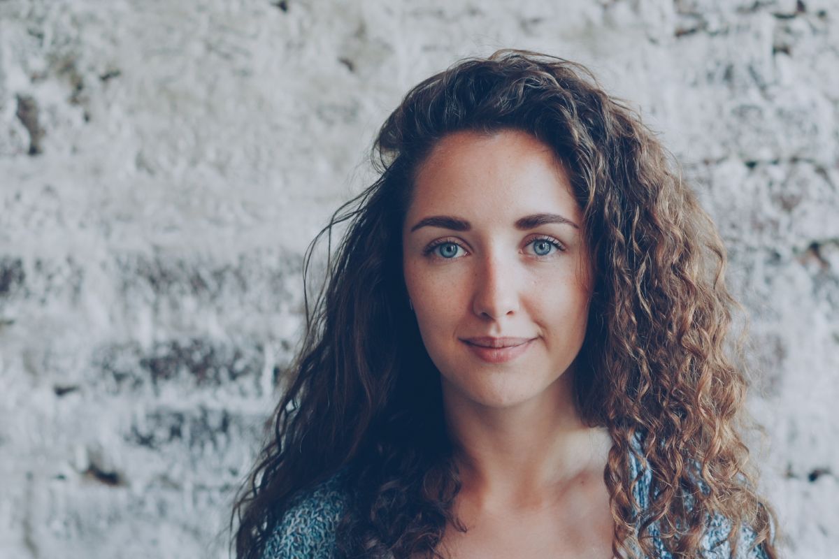 Young woman with curly brown hair standing against a white painted brick wall.