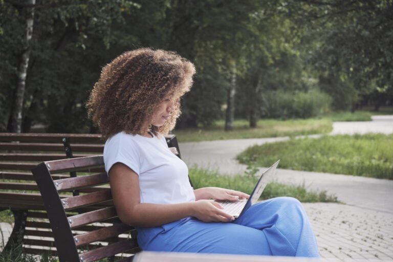 Young African American woman sitting on park bench working on her laptop