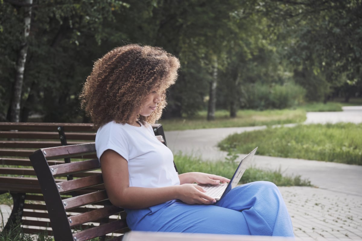 Young African American woman sitting on park bench working on her laptop